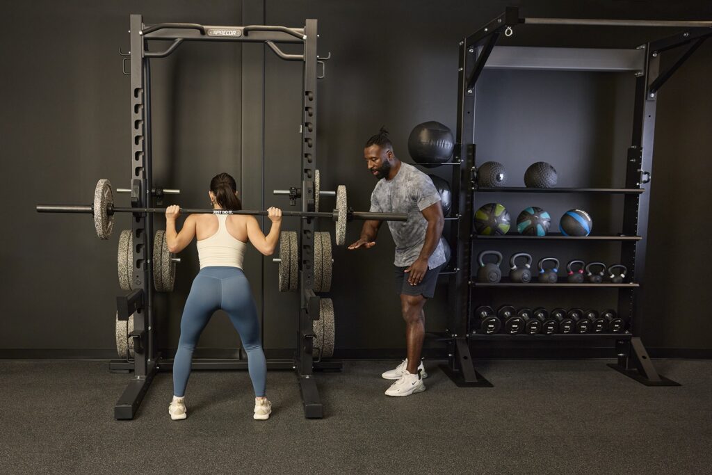 A trainer spots a woman doing squats with a Nike half rack.