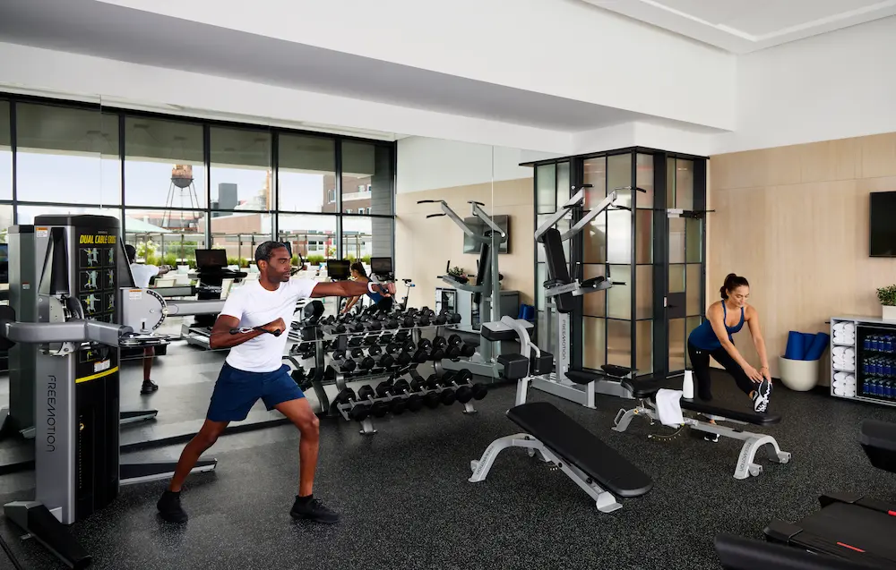 A gym interior with a man exercising and a woman stretching, surrounded by various fitness equipment and large windows.