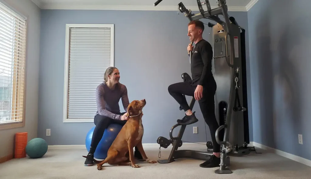 A man exercises with gym equipment while a woman pets a dog nearby in a home gym setting.